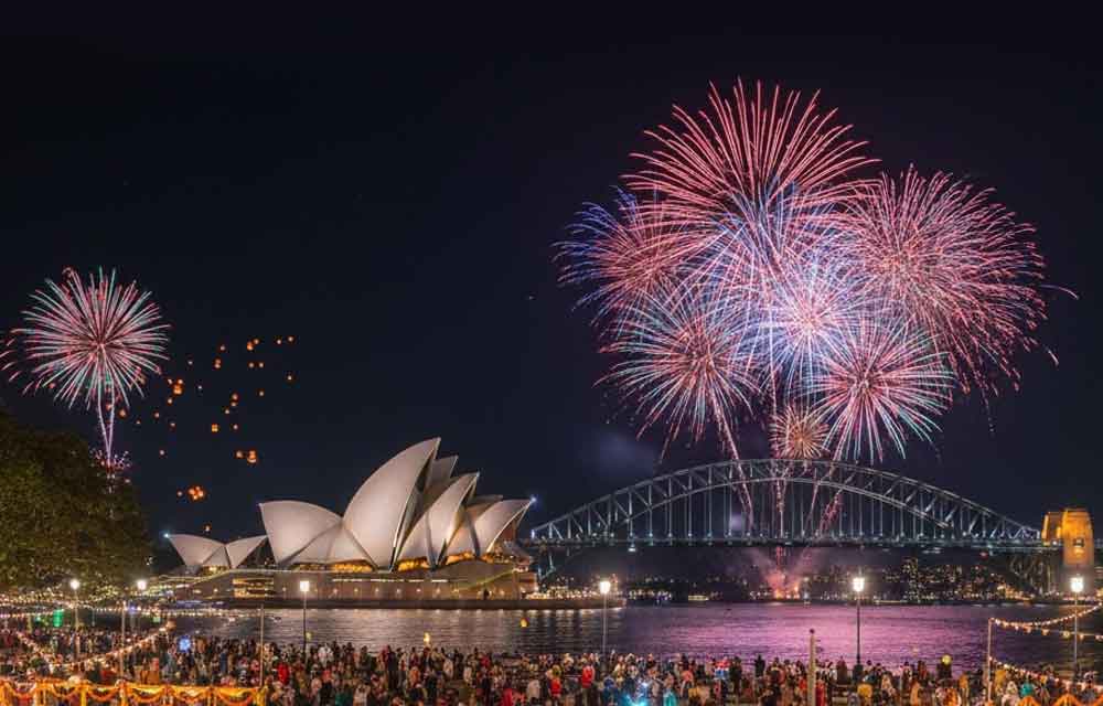 A festive depiction of Diwali 2025 celebrations in Australia. In the foreground, a diverse crowd of people in traditional Indian attire gather on a grassy area, many holding sparklers and diyas, with colorful rangoli visible. In the background, the iconic Sydney Opera House and Sydney Harbour Bridge are illuminated against a night sky filled with vibrant fireworks. A "Diwali 2025" calendar icon is in the bottom right corner.