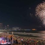 An aerial nighttime view of a vibrant Diwali celebration on a Durban beach. A large crowd of people in traditional Indian attire (sarees and kurtas) gathers around a gigantic, illuminated Om symbol made of hundreds of lit earthen lamps (diyas) placed on the sand.
