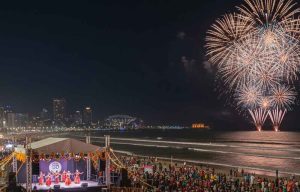 An aerial nighttime view of a vibrant Diwali celebration on a Durban beach. A large crowd of people in traditional Indian attire (sarees and kurtas) gathers around a gigantic, illuminated Om symbol made of hundreds of lit earthen lamps (diyas) placed on the sand.