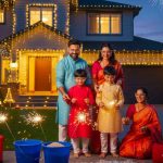 A smiling family of four, dressed in traditional Indian attire, celebrating Diwali safely in front of their house. The father and two young sons are holding sparklers, while the mother kneels nearby. Safety equipment like buckets of water/sand, a fire extinguisher, and a first aid kit are prominently displayed around a colorful rangoli, with the house adorned in festive lights and fireworks in the night sky