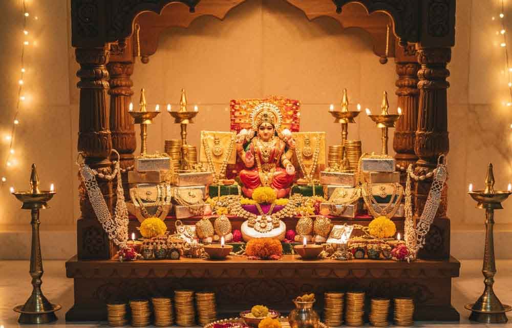 A close-up view of a Dhanteras puja altar featuring gold and silver coins, jewelry, and metal utensils, surrounded by lit clay diyas and festive offerings like flowers and sweets.