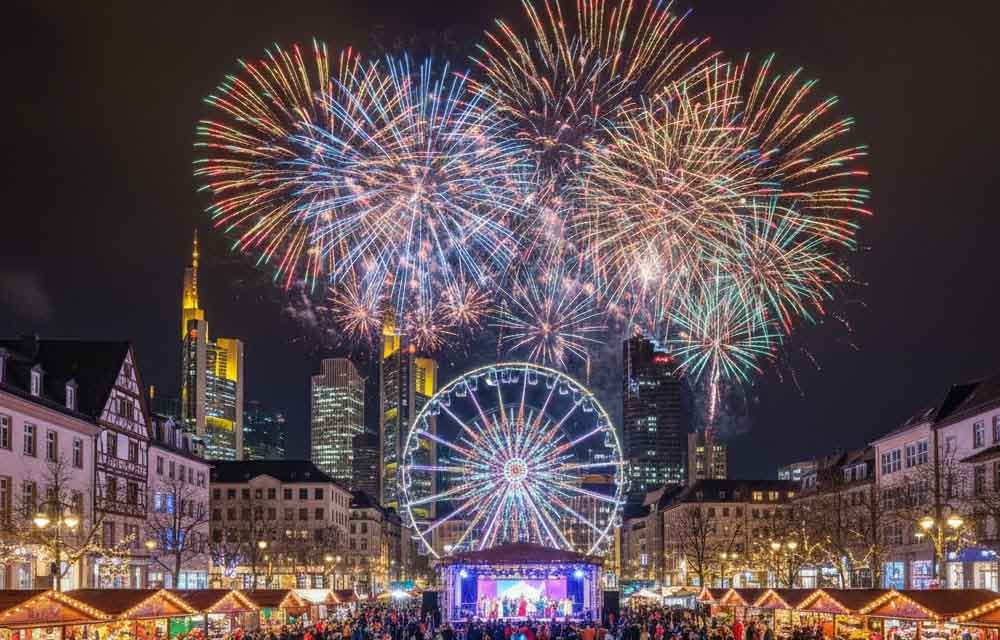 Diwali festival celebration at night in Germany, featuring fireworks over a city and a large gathering with people dressed in festive attire.