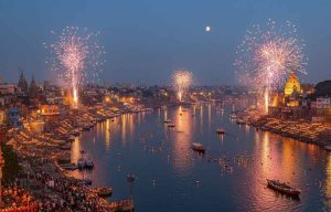 An expansive view of Dev Deepawali in Varanasi at dusk, with thousands of illuminated diyas lining the ghats of the Ganges River, numerous boats adorned with lights, and colorful fireworks exploding over the cityscape under a crescent moon.