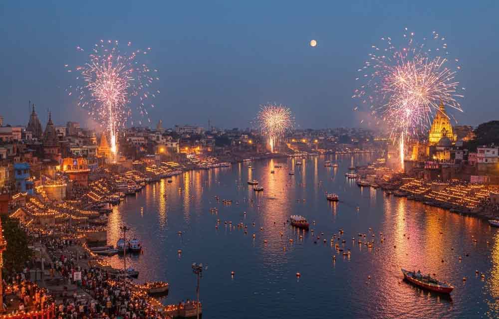 An expansive view of Dev Deepawali in Varanasi at dusk, with thousands of illuminated diyas lining the ghats of the Ganges River, numerous boats adorned with lights, and colorful fireworks exploding over the cityscape under a crescent moon.