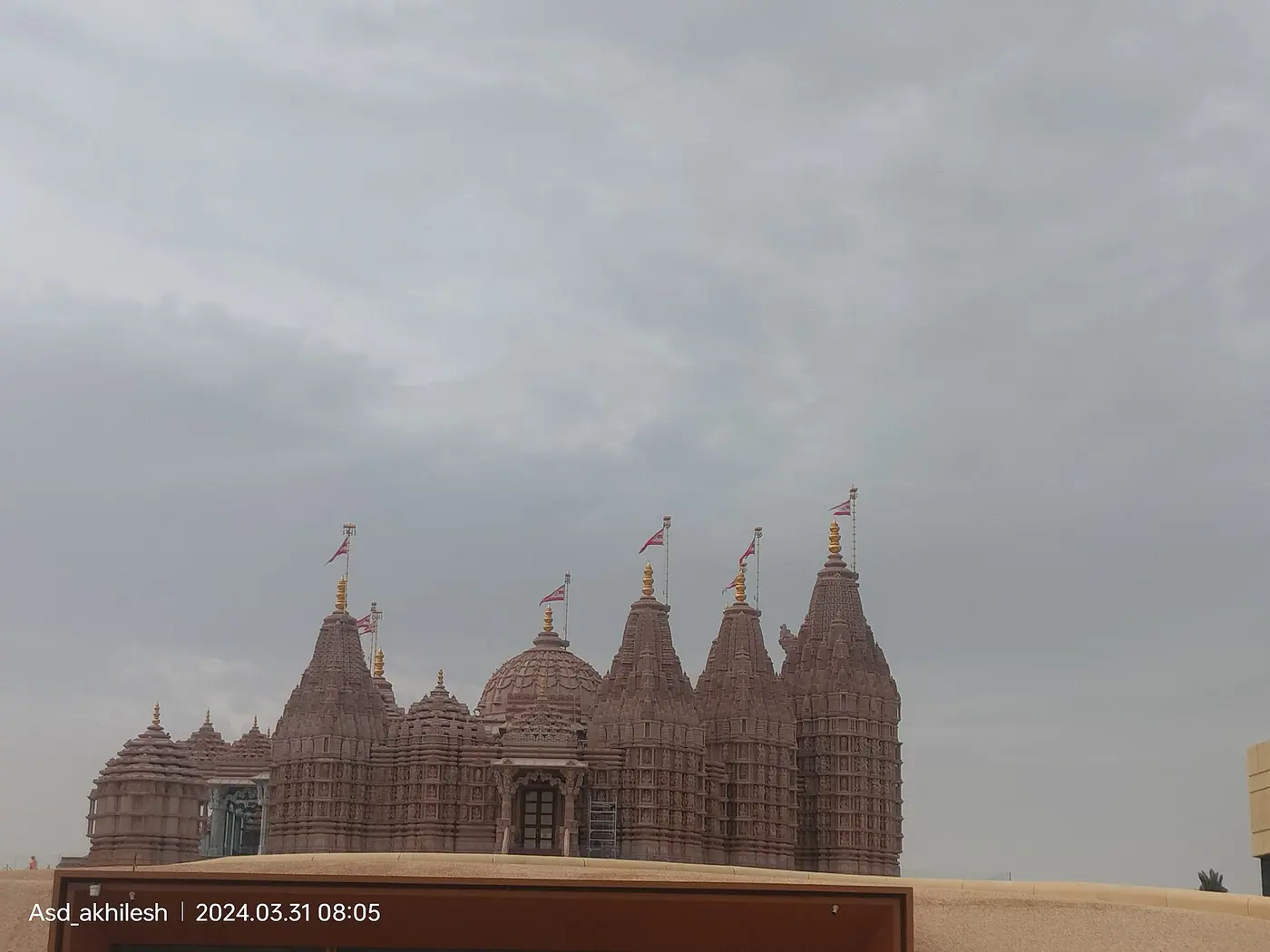 Pink sandstone spires and dome of the BAPS Hindu Mandir in Abu Dhabi, with saffron flags, seen over a sand embankment under an overcast sky